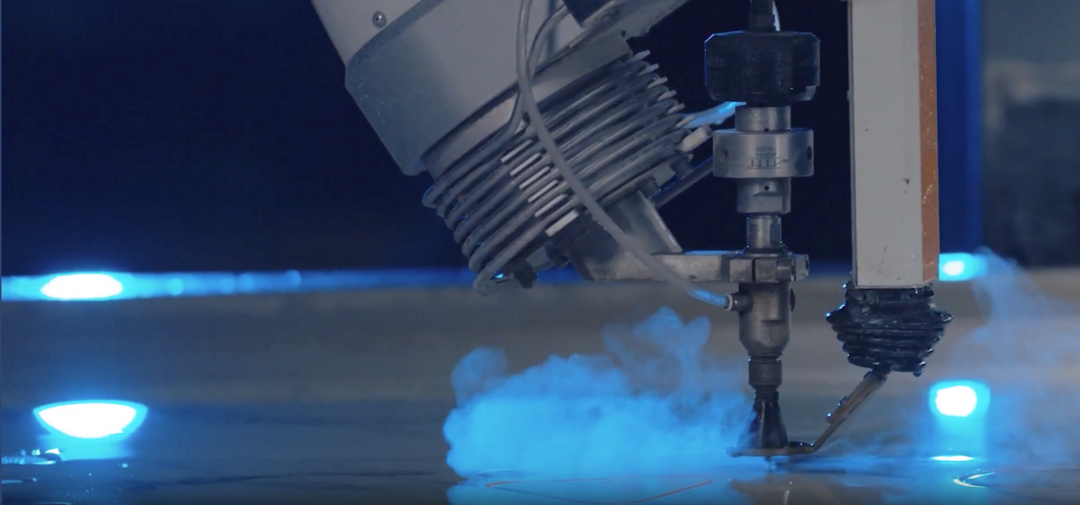Ein Wasserstrahlschneider schneidet präzise durch ein Material, während Dampf aufsteigt umgeben von blauer Beleuchtung, in einer industriellen Umgebung.