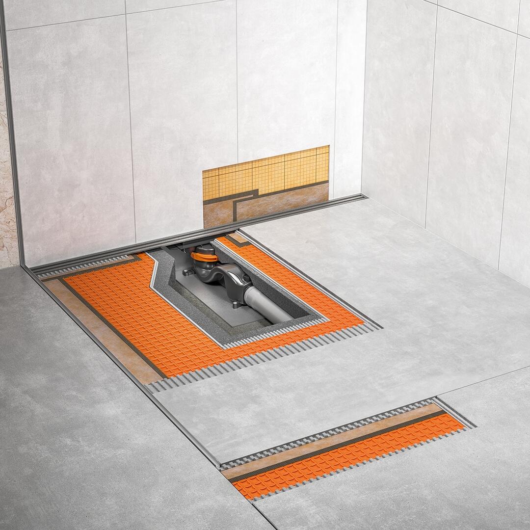 Eine moderne, grau geflieste Duschfläche in einer Ecke. In den Boden eingelassen und noch nicht überfliest sind flache, orange Abflusssysteme.