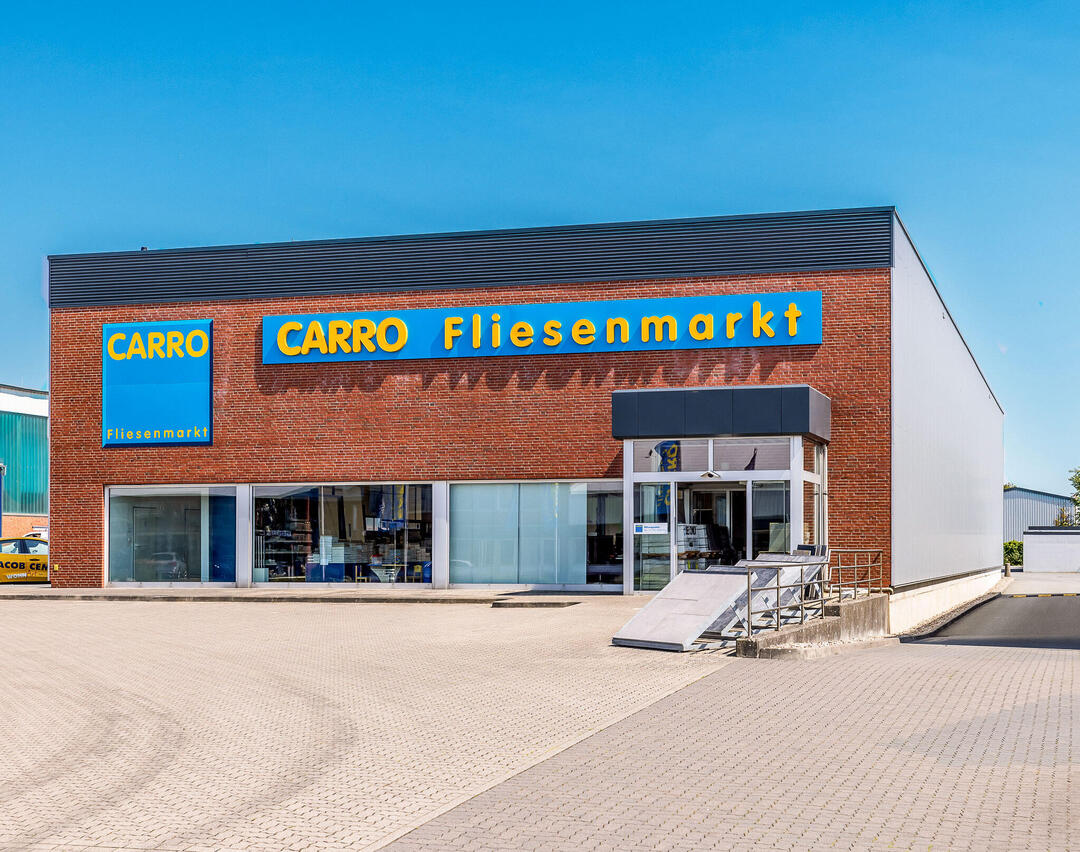 Der CARRO Fliesenmarkt in Husum schräg von vorne fotografiert unter strahlend blauem Himmel: Ein rechteckiges Backsteingebäude mit einem gepflasterten Vorplatz.