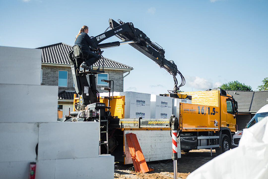 Ein Lastwagen mit Kran entlädt weiße Baustoffblöcke auf einer Baustelle. Ein Arbeiter steuert den Kran. Im Hintergrund ist ein modernes Wohnhaus unter einem blauen Himmel zu sehen.