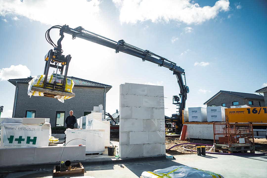Ein Kran hebt Baumaterialien auf einer Baustelle, während ein Arbeiter daneben steht. Im Hintergrund sind teilweise errichtete Gebäude und Stapel von Baumaterialien sichtbar.
