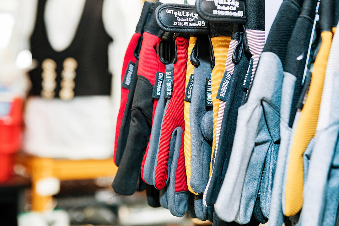 Mehrere Arbeitshandschuhe hängen nebeneinander an einem Verkaufsstand. Im Hintergrund ist ein schwarz-weißes Kleidungsstück sichtbar, verschwommen. Handschuhe sind in verschiedenen Farben, wie Rot und Grau, dargestellt.