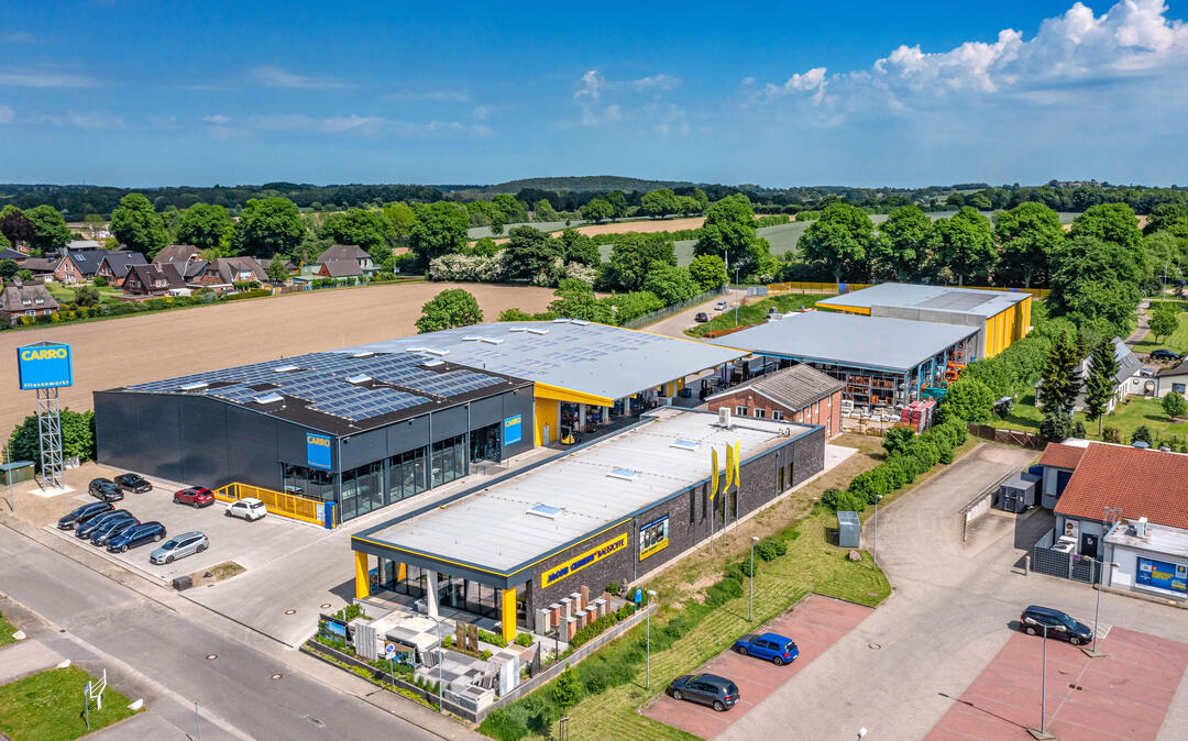 Der CARRO Fliesenmarkt in Bad Segeberg aus der Luft fotografiert: Ein großzügiger Gebäudekomplex, der von Bäumen umgeben ist, unter blauem Himmel.