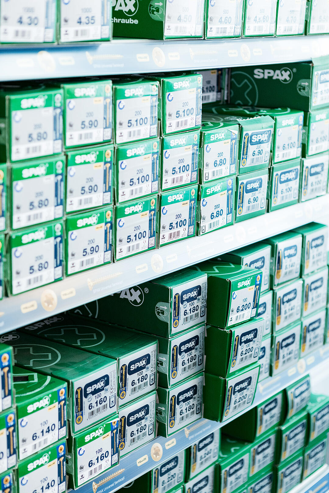 Regale mit grün-weißen Verpackungen von SPAX-Schrauben, ordentlich gestapelt. Jedes Paket ist mit einer Zahl und Information wie '5.10', '6.80' gekennzeichnet.