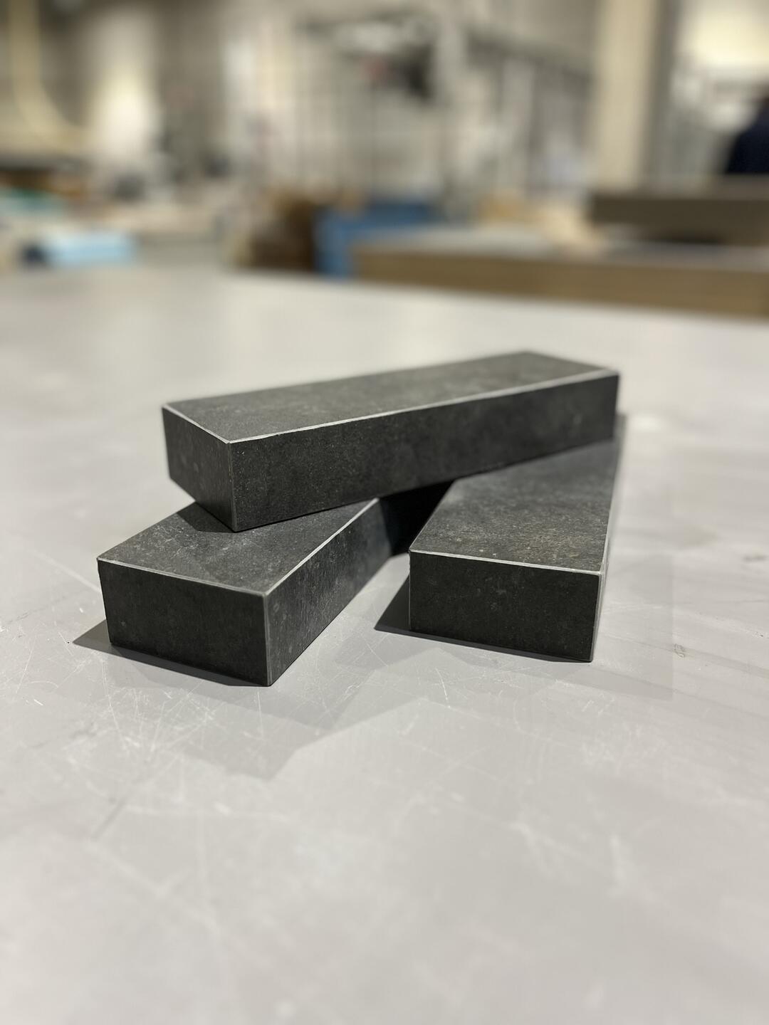 Drei schwarze rechteckige Blöcke liegen übereinander auf einer glatten, grauen Oberfläche in einem unscharfen industriellen Raum.