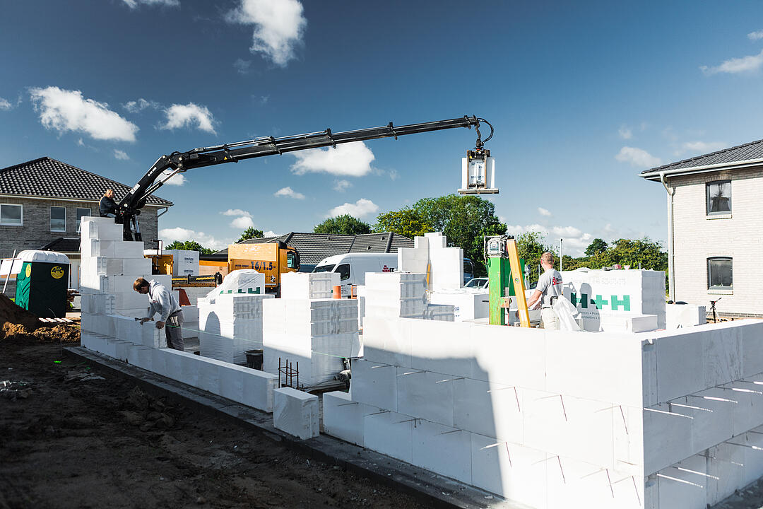 Ein Kran hebt in einer Vorstadtumgebung unter blauem Himmel mit Wolken einen Betonblock auf eine teilweise errichtete Mauer, während Arbeiter auf der Baustelle tätig sind.