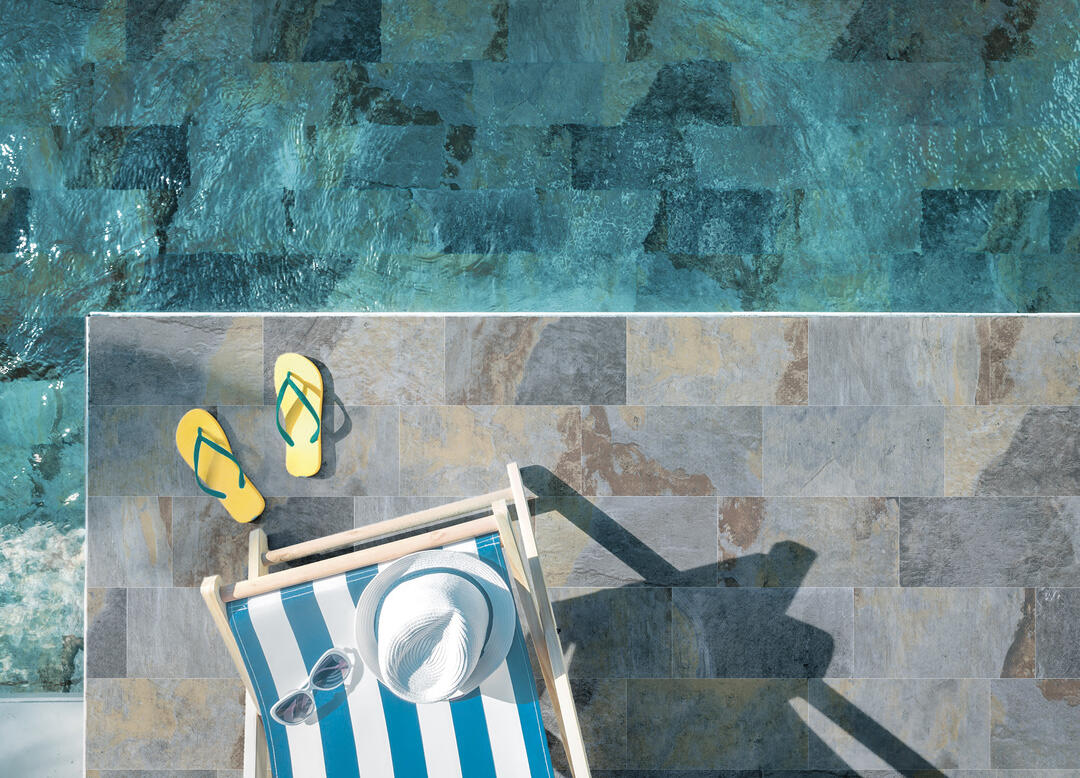 Luftaufnahme eines Pools mit türkisen Fliesen, davor gelbe Flip-Flops, ein blau-weiß gestreifter Liegestuhl mit Sonnenhut und Sonnenbrille auf grauem Steinboden