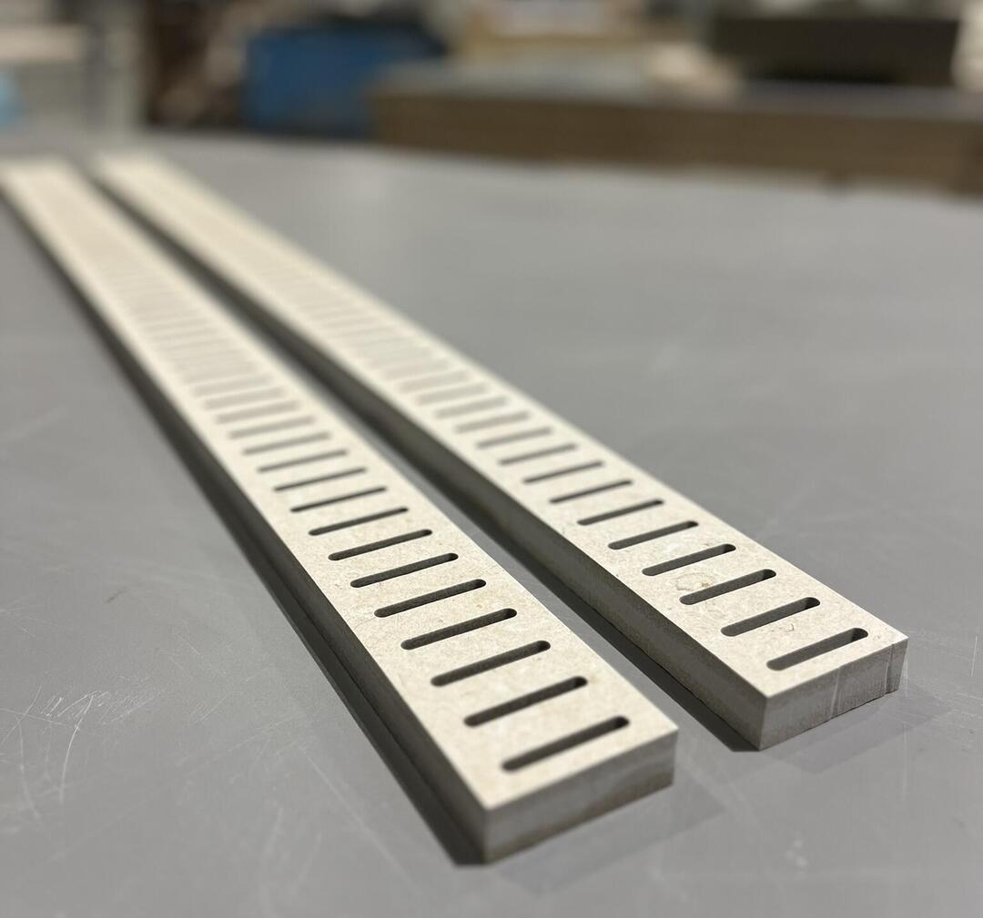 Zwei graue, schmale Metallstreifen mit gleichmäßigen Schlitzen liegen nebeneinander auf einer grauen Tischoberfläche in einer industriellen Umgebung.