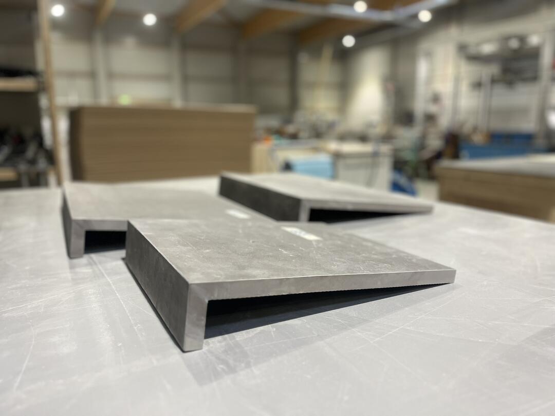 Metallplatten liegen auf einer Tischoberfläche. Sie sind rechtwinklig gebogen und scheinen massiv zu sein. Im Hintergrund ist eine Werkstatt zu sehen.