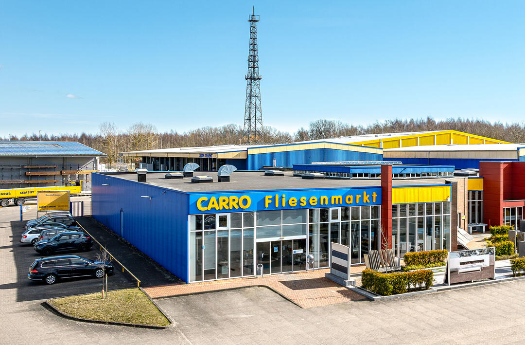 Der CARRO Fliesenmarkt in Greifswald, aus halber Höhe von einer Drohne fotografiert: Ein modernes Gebäude mit großen Fensterfronten und dem blauen CARRO-Schriftzug, unter blauem Himmel.