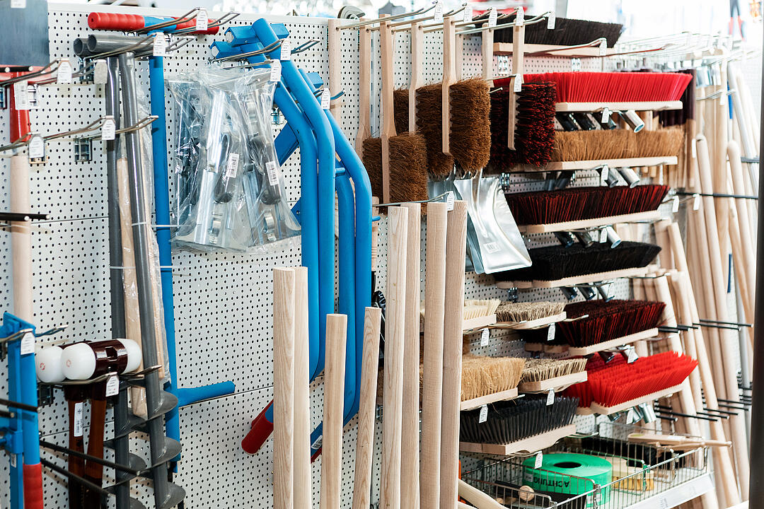 Verschiedene Handwerkzeuge hängen und stehen ordentlich auf einem weißen, perforierten Wandregal in einem Laden: Besen, Kehrbleche, blaue Metallsägen und Holzstiele.