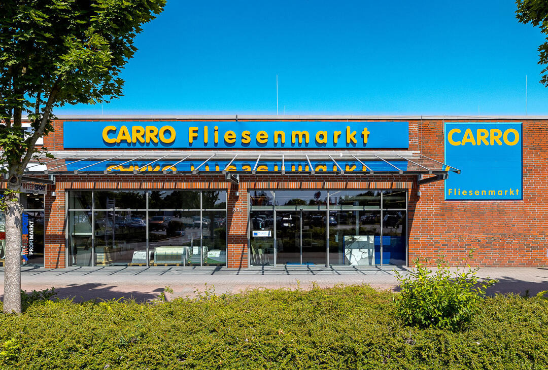 Der CARRO Fliesenmarkt in Rendsburg, von vorne fotografiert: Ein flaches rotes Backsteingebäude mit blauem CARRO-Schriftzug und Fensterfront unter blauem Himmel mit Bäumen und Hecken im Vordergrund.