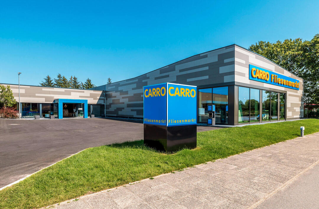 Der CARRO Fliesenmarkt in Flensburg, ein graues Rechteck, schräg von vorne fotografiert, mit dem Parkplatz und einem blauen CARRO-Quader im Vordergrund, unter blauem Himmel.