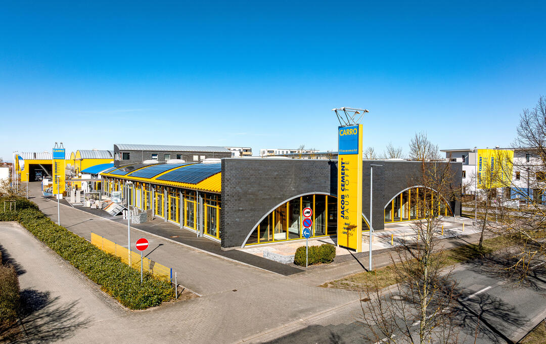 Der CARRO Fliesenmarkt in Neubrandenburg, von einer Drohne aus halber Höhe fotografiert: Ein moderner Gebäudekomplex mit einem großen gelben Turm mit dem Unternehmensschriftzug im Vordergrund unter blauem Himmel. Auf diesem Bild sind auch Bäume zu sehen.