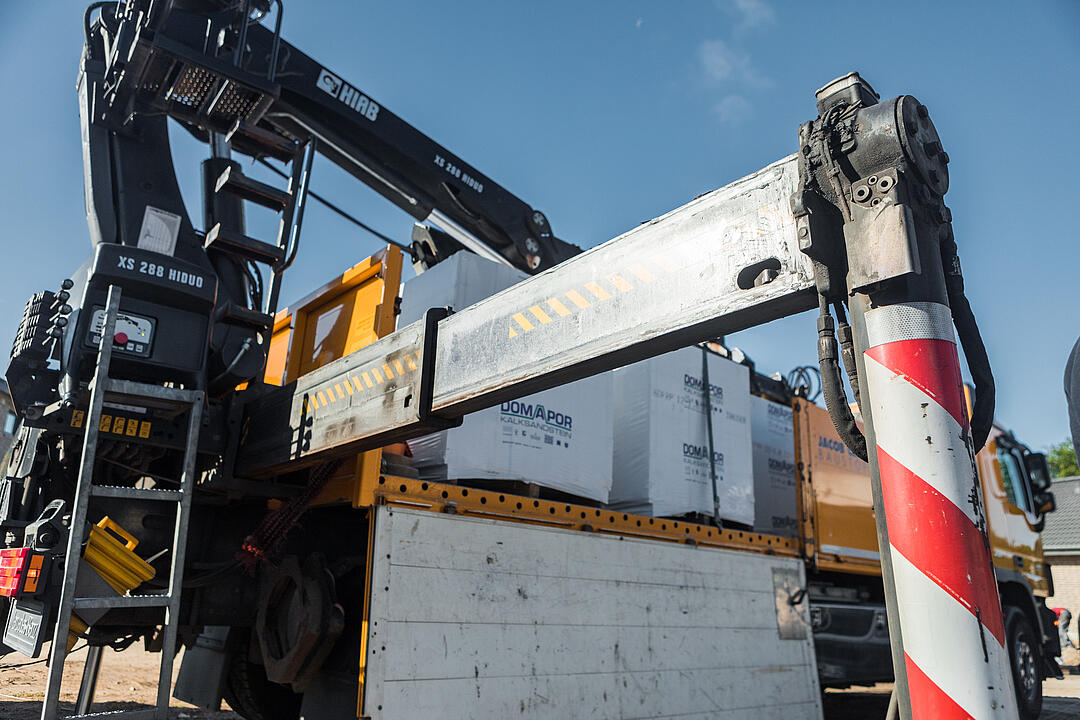 Ein Lastwagen mit Kranarm hebt Baumaterialien, während der Lastwagen auf einer Baustelle steht, umgeben von klarem Himmel.