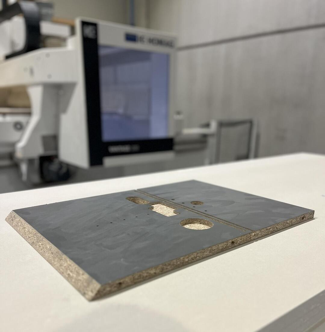 Eine rechteckige, graue Platte mit Aussparungen liegt auf einem weißen Tisch. Im Hintergrund befindet sich eine industrielle Maschine in einer Werkstattumgebung.