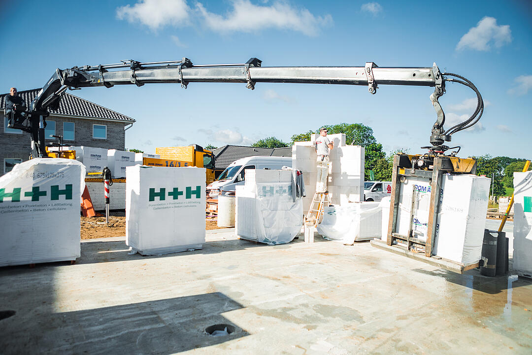 Ein Baukran hebt große, weiße H+H-Materialblöcke auf einer Baustelle an. Im Hintergrund sind weitere Baugeräte, ein Haus und grüne Bäume zu sehen.